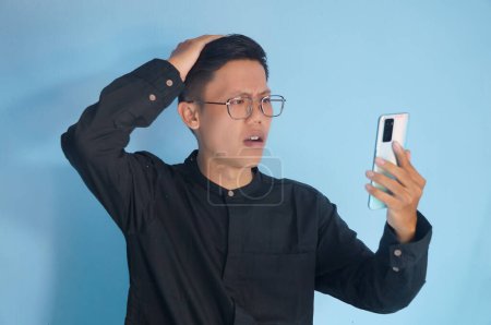 Joven asiático hombre mostrando confusa expresión facial mientras sostiene el teléfono móvil
