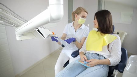 Photo for High-qualified dentist shows the young female patient an X-ray of the teeth in the dental chair at modern clinic. Explains and explores x-ray treatment. - Royalty Free Image