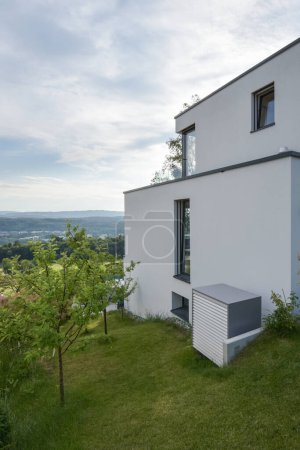 Téléchargez les photos : Unité externe d'une pompe à chaleur à air devant une maison moderne basse énergie en blanc - en image libre de droit
