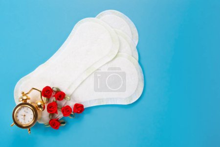 Wecker, Blumen und feminine Hygienepads auf blauem Hintergrund. Konzept der ersten Menstruationsperiode, Zykluszeit