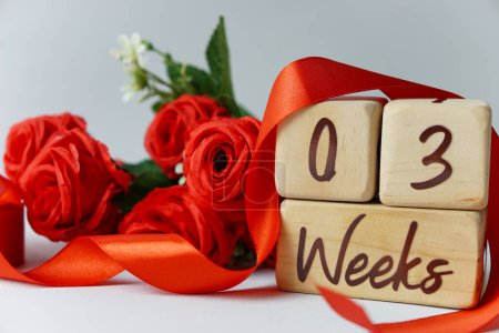 Téléchargez les photos : Jalon de l'âge gestationnel de 3 semaines écrit sur un cube en bois avec des roses rouges et des rubans, et un fond blanc - en image libre de droit