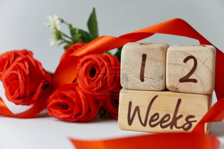 Téléchargez les photos : Jalon de l'âge gestationnel de 12 semaines écrit sur un cube en bois avec des roses rouges et des rubans, et un fond blanc - en image libre de droit