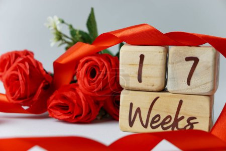 Téléchargez les photos : 17 semaines d'âge gestationnel jalon écrit sur un cube en bois avec des roses rouges et des rubans, et un fond blanc - en image libre de droit