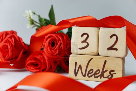 Téléchargez les photos : 32 semaines d'âge gestationnel jalon écrit sur un cube en bois avec des roses rouges et des rubans, et un fond blanc - en image libre de droit