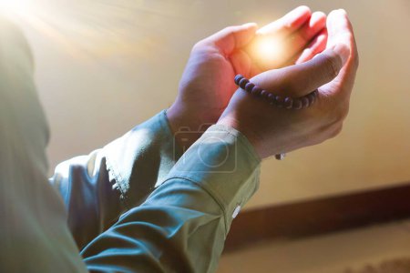 the hands of a man in Muslim prayer holding prayer beads, with light between his palms