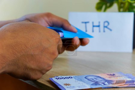 A portrait of a hand putting rupiah notes into an envelope for THR. THR or Tunjangan Hari Raya is a holiday allowance or bonus given ahead of Ramadan.