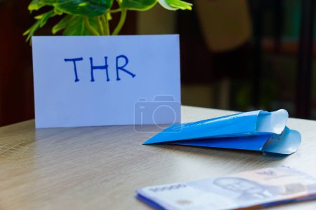 potret uang kertas rupiah dan amplop biru untuk THR di atas meja.THR or Tunjangan Hari Raya is a holiday allowance or bonus given ahead of Ramadan.