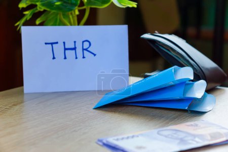 potret uang kertas rupiah dan amplop biru untuk THR di atas meja.THR oder Tunjangan Hari Raya ist ein Urlaubsgeld oder Bonus, der vor dem Ramadan gewährt wird.