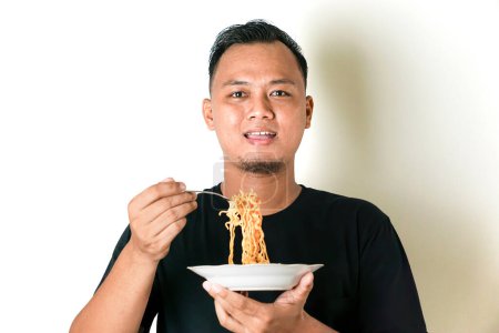 A man wearing a black shirt is holding a plate of noodles and eating with a fork. He looks content and is posing against a white background