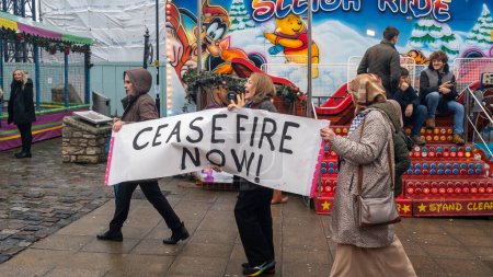 Photo for Protest walk calling for Cease fire, Southampton, UK,  16 December, 2023.Three woman are marching with a banner that reads "CEASE FIRE NOW!" in bold letters, passing by a colorful amusement park ride. - Royalty Free Image