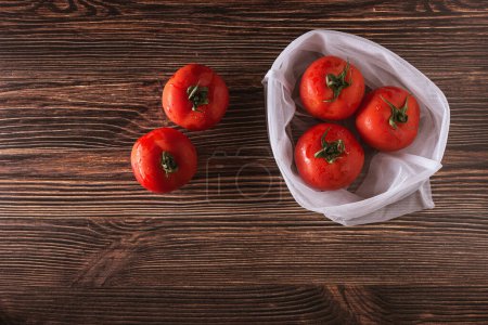 Photo for Mesh bag made of ecological material with fresh tomatoes inside and outside on a dark brown wooden table. - Royalty Free Image