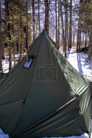 Dieses Bild zeigt ein Tipi-Zelt, das in einer verschneiten Landschaft aufgestellt wurde, mit einem aus dem Zelt ragenden Metallschornstein, der auf einen gemütlichen Zeltplatz inmitten der winterlichen Wildnis hindeutet..