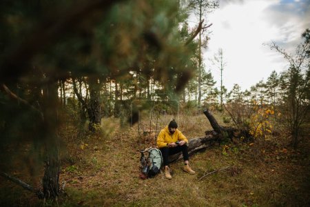 Foto de Un hombre barbudo descansa sobre un árbol caído en medio del bosque. Un hombre usa un teléfono móvil para buscar GPS. - Imagen libre de derechos