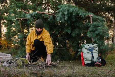 Survival in the wild. A bearded man lights a fire near a makeshift shelter made of pine branches.