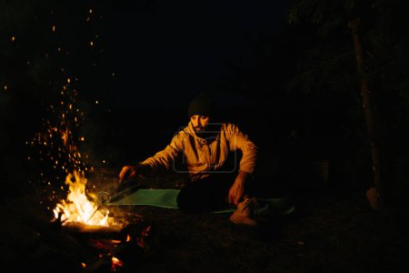 Foto de El viajero descansa junto a la fogata nocturna. Amueblado refugio de ramas para dormir con una lámpara de queroseno y un fuego rodeado de piedras. - Imagen libre de derechos