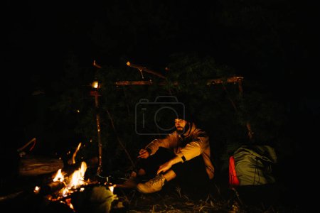 Foto de El viajero descansa junto a la fogata nocturna. Amueblado refugio de ramas para dormir con una lámpara de queroseno y un fuego rodeado de piedras. - Imagen libre de derechos