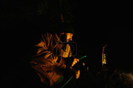 Foto de Un viajero descansa junto a una fogata nocturna con un teléfono en sus manos. Un hombre revisa su teléfono, noticias, mensajes. - Imagen libre de derechos