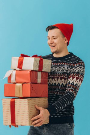 Photo for Young man in Christmas sweater and red hat, hold many present boxes with gift ribbon bow isolated on blue background. Happy New Year, celebration concept - Royalty Free Image