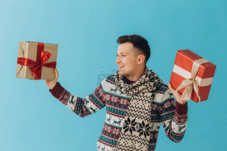 Foto de Hombre joven en jersey de Navidad y bufanda que sostiene muchas cajas de regalo con lazo de cinta de regalo aislado sobre fondo azul. Feliz año nuevo, concepto de celebración. - Imagen libre de derechos