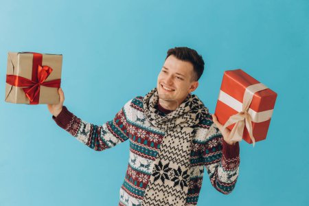 Téléchargez les photos : Jeune homme en pull de Noël et écharpe tenant de nombreuses boîtes-cadeaux avec ruban cadeau arc isolé sur fond bleu. Bonne année, concept de célébration. - en image libre de droit