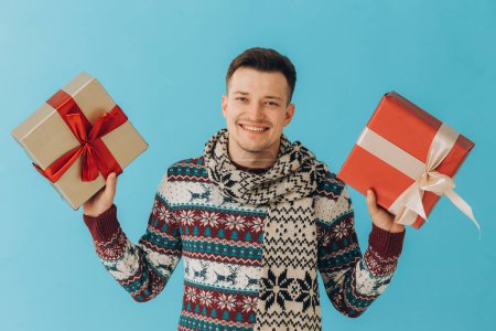 Photo for Young man in Christmas sweater and scarf holding many gift boxes with gift ribbon bow isolated on blue background. Happy new year, celebration concept. - Royalty Free Image