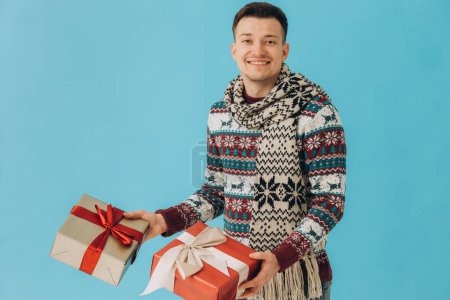 Photo for Young man in Christmas sweater and scarf holding many gift boxes with gift ribbon bow isolated on blue background. Happy new year, celebration concept. - Royalty Free Image