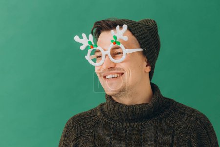 Foto de Un joven con un suéter cálido, una gorra y unas gafas de carnaval aisladas sobre un fondo verde. - Imagen libre de derechos