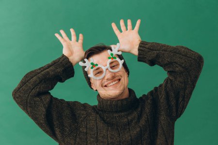 Téléchargez les photos : Un jeune homme dans un pull chaud, une casquette et des lunettes de carnaval isolés sur un fond vert. - en image libre de droit