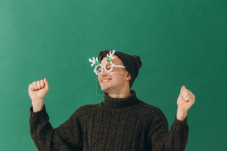 Foto de Un joven con un suéter cálido, una gorra y unas gafas de carnaval aisladas sobre un fondo verde. - Imagen libre de derechos
