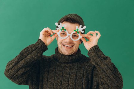 Photo for A young man in a warm sweater, a cap and carnival glasses isolated on a green background. - Royalty Free Image
