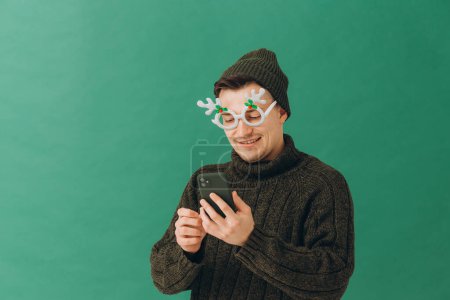 Foto de Un joven con un suéter cálido, gafas de carnaval y un teléfono en las manos, aislado sobre un fondo verde. - Imagen libre de derechos