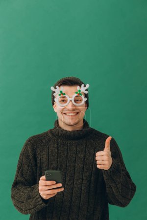 Téléchargez les photos : Un jeune homme dans un pull chaud, lunettes de carnaval et un téléphone dans ses mains, isolé sur un fond vert. - en image libre de droit