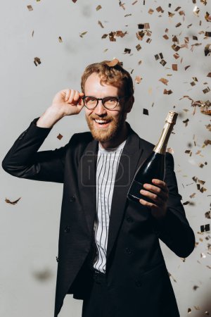 Foto de Celebración del nuevo año. Joven pelirrojo con una botella de champán aislado sobre un fondo blanco bajo confeti. - Imagen libre de derechos