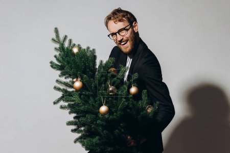 Foto de Hombre guapo de pelo rojo con una chaqueta negra y gafas baila con un árbol de Navidad en sus manos. concepto de fiesta de Año Nuevo. - Imagen libre de derechos