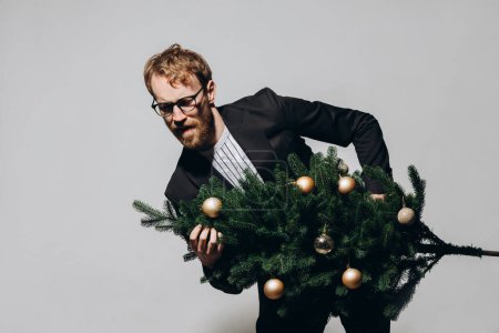 Téléchargez les photos : Un bel homme roux vêtu d'une veste noire et portant des lunettes danse avec un sapin de Noël à la main. Concept de fête Nouvel An. - en image libre de droit