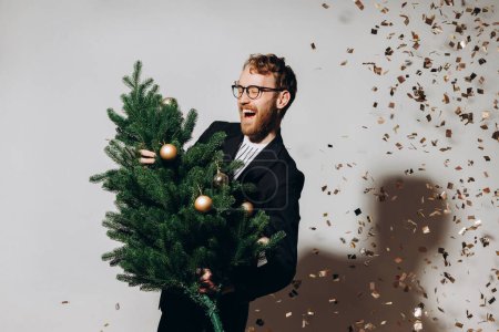 Photo for Red-haired handsome man in a black jacket and glasses dances with a Christmas tree in his hands. New Year party concept. - Royalty Free Image