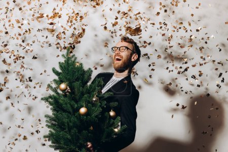 Téléchargez les photos : Un bel homme roux vêtu d'une veste noire et portant des lunettes danse avec un sapin de Noël à la main. Concept de fête Nouvel An. - en image libre de droit