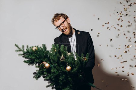Foto de Hombre guapo de pelo rojo con una chaqueta negra y gafas baila con un árbol de Navidad en sus manos. concepto de fiesta de Año Nuevo. - Imagen libre de derechos