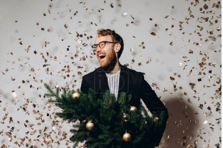 Photo for Red-haired handsome man in a black jacket and glasses dances with a Christmas tree in his hands. New Year party concept. - Royalty Free Image