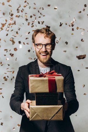 Foto de Retrato de un joven feliz con una gran sonrisa sosteniendo regalos mientras caen los confeti dorados - Imagen libre de derechos