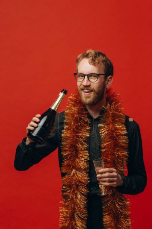 Téléchargez les photos : Un bel homme aux cheveux roux dans des verres et un boa doré lève une coupe de champagne. Célébration du nouvel an. - en image libre de droit