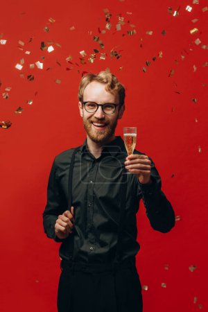 Foto de Concepto de Navidad, fiesta y vacaciones. Hombre guapo levantando una copa de champán y sonriendo bajo el confeti sobre fondo rojo. - Imagen libre de derechos
