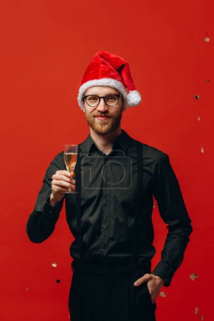 Téléchargez les photos : Noël, fête et concept de vacances. Charmant homme au chapeau de Père Noël levant un verre de champagne et souriant, célébrant le Nouvel An, fond rouge. - en image libre de droit