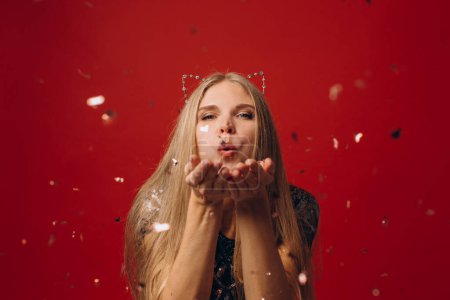 Foto de Foto de mujer bonita alegre soplando confeti de oro de manos aisladas sobre fondo rojo - Imagen libre de derechos