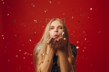 Foto de Foto de alegre encantadora hermosa chica soplando confeti de sus manos aisladas sobre fondo rojo. - Imagen libre de derechos