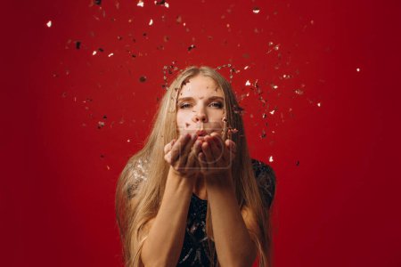 Téléchargez les photos : Photo de joyeuse charmante belle fille soufflant des confettis de ses mains isolées sur fond rouge. - en image libre de droit