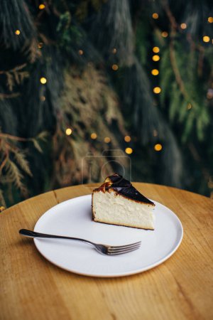 Foto de Un delicioso trozo de pastel de queso o pastel en la mesa. Fondo de ramas de pino y guirnaldas festivas. - Imagen libre de derechos