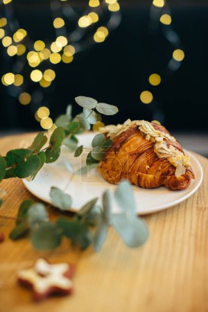Foto de Un delicioso croissant sobre una mesa en una cafetería decorada con luces navideñas festivas. - Imagen libre de derechos