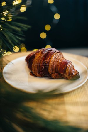Foto de Un delicioso croissant sobre una mesa en una cafetería decorada con luces navideñas festivas. - Imagen libre de derechos