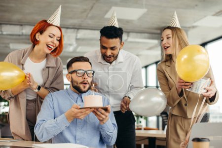 Foto de Feliz hombre de negocios soplando una vela en la torta mientras celebra el cumpleaños con compañeras de trabajo en la oficina. - Imagen libre de derechos
